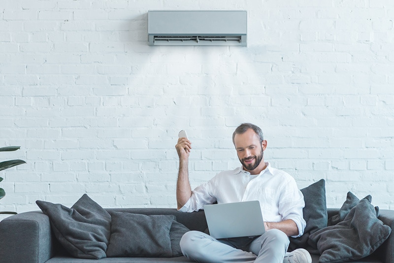 Man sitting on couch using laptop under ductless heating and cooling vent.