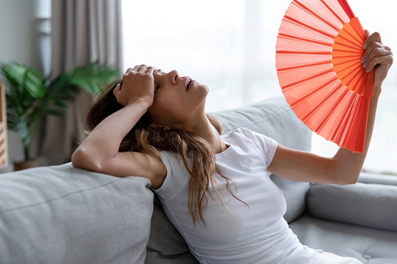 Why Is My AC Blowing Hot Air? Photo of an overheated woman fanning herself in her living room.