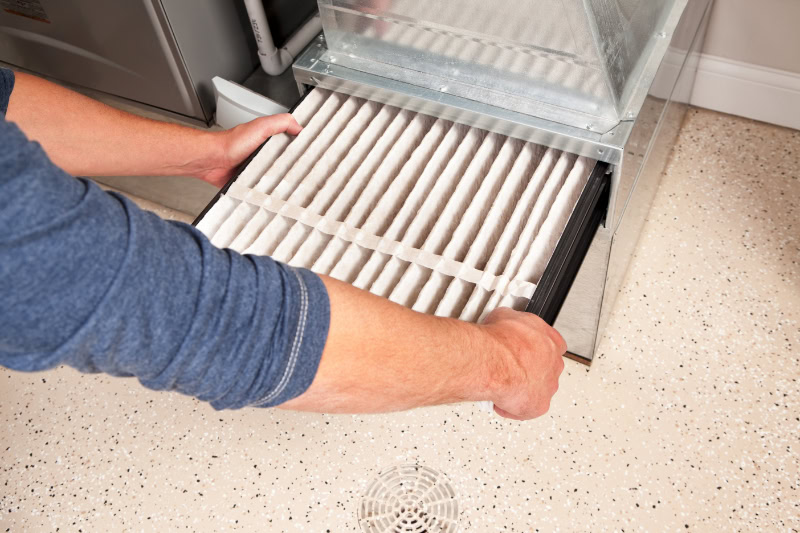 HVAC technician installing a furnace air filter. This filter is not brand new but it is relatively clean, it was removed for inspection. This would be a typical filter style found in a high end newer house. The floor is epoxy painted concrete with traction chips and a floor drain.